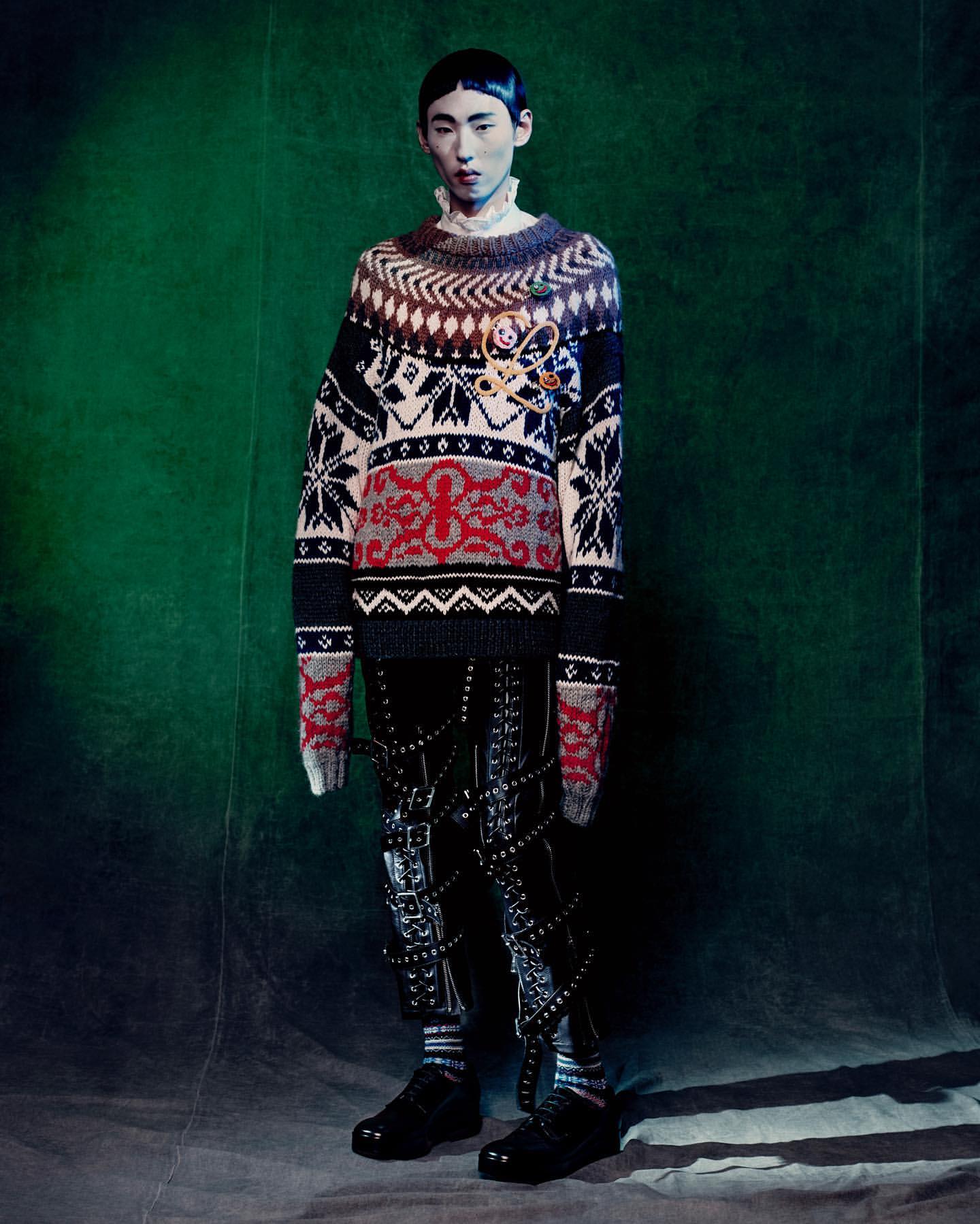 Asian model stands in a photo studio wearing a patterned sweater, black pants with straps, and black shoes against a green backdrop.