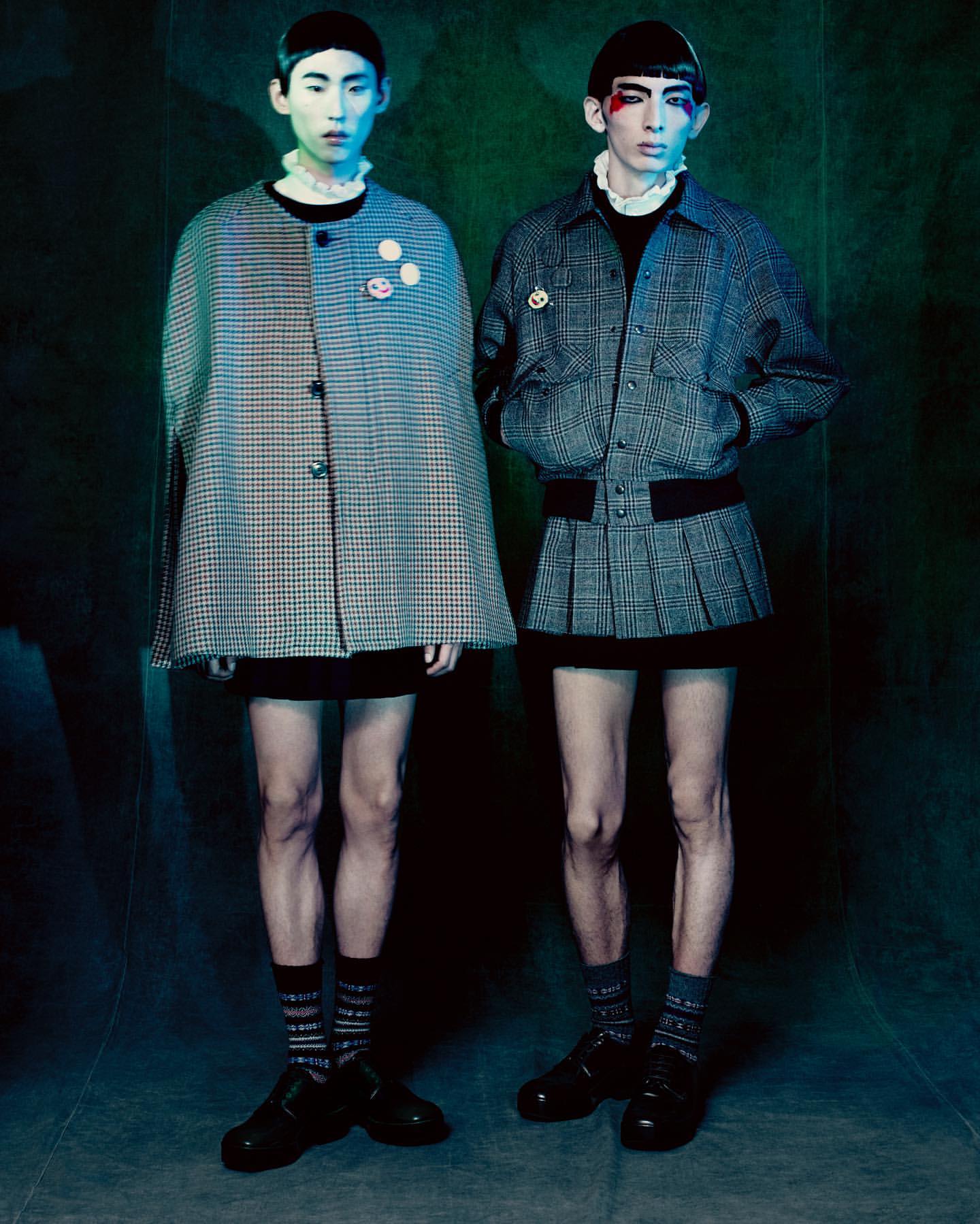 Two young adult fashion models in vintage-inspired outfits with pale complexions, posed indoors against a blue-green backdrop with soft lighting.