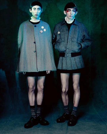 Two young adult fashion models in vintage-inspired outfits with pale complexions, posed indoors against a blue-green backdrop with soft lighting.