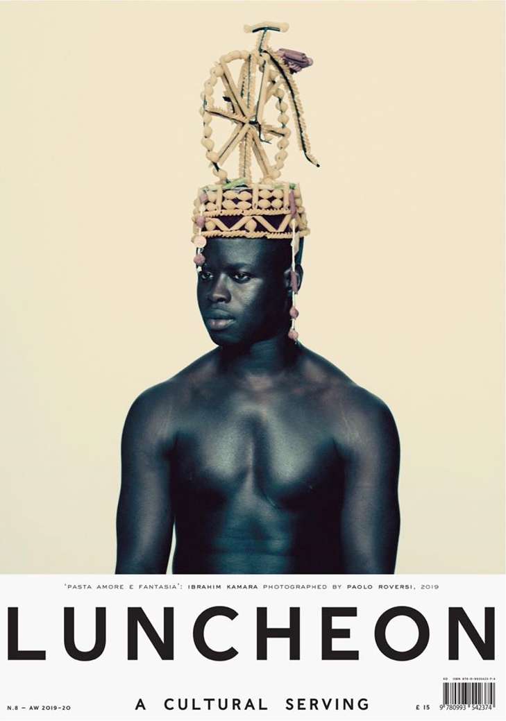 Striking editorial portrait of a shirtless Black man wearing a decorative beaded headpiece with a bicycle on top, set against a light beige backdrop.