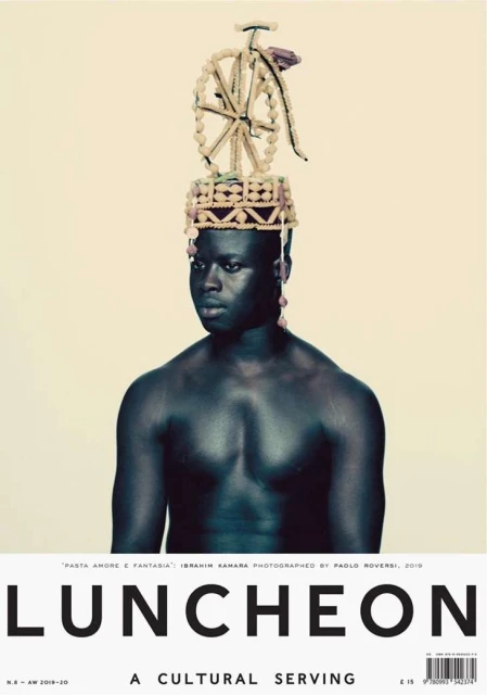 Striking editorial portrait of a shirtless Black man wearing a decorative beaded headpiece with a bicycle on top, set against a light beige backdrop.