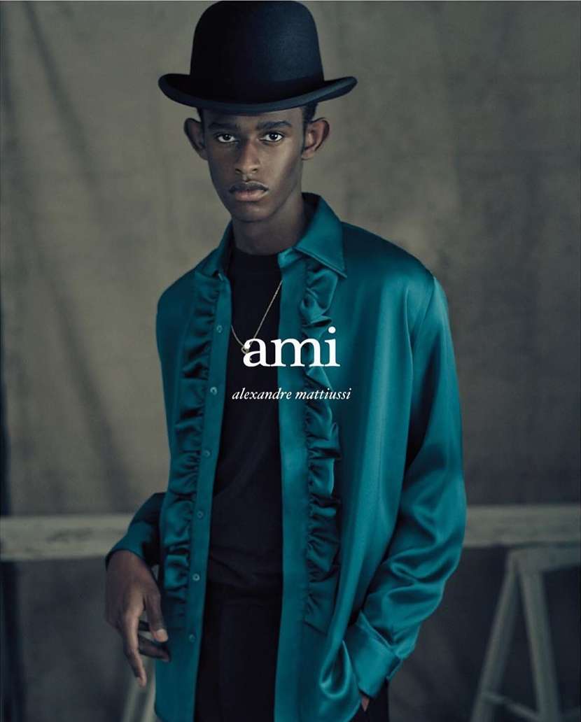 Portrait of a black male model wearing a black bowler hat and teal, ruffled, long-sleeved shirt with logo and a gold chain necklace.