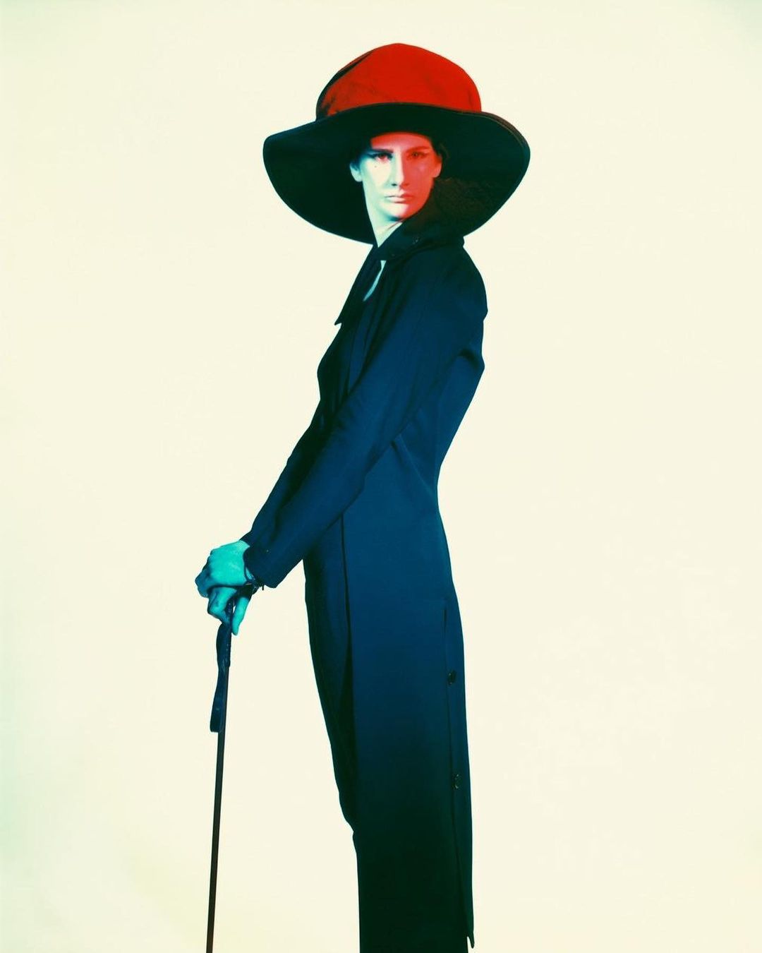 Fashionable woman in a black overcoat and wide-brimmed hat posing indoors, holding a cane, against a pale neutral backdrop.