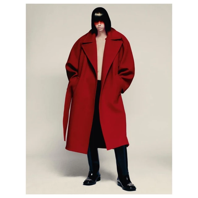 A person with short black hair and red visor stands in a studio wearing a red oversized coat, black pants, and black boots.