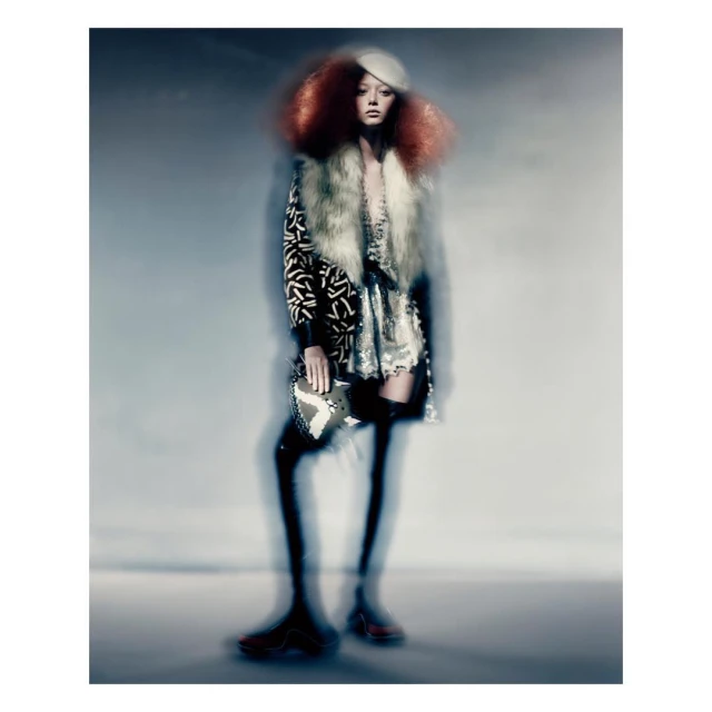 Full-length fashion shot with a blurry effect showcasing a model in an ornate coat, thigh-high stockings, headdress, and statement accessories.
