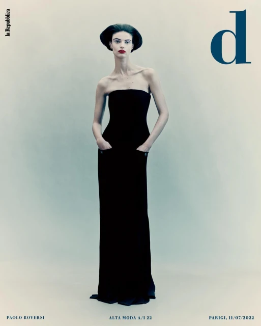Editorial fashion shot of a tall, thin, adult female model in a black strapless gown with hands in pockets, against a solid light background.