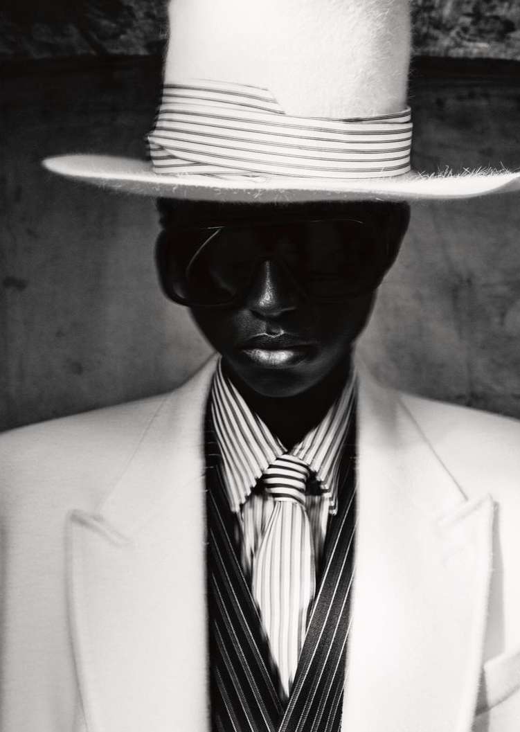 Elegant studio portrait of a Black person wearing a white suit, striped shirt and tie, sunglasses, and a white striped top hat.