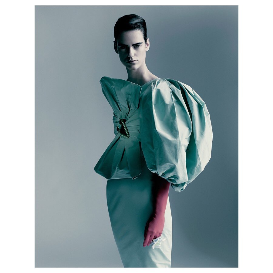 A woman poses in a teal high-fashion gown with a large bow detail and pink gloves against a light grey studio backdrop.