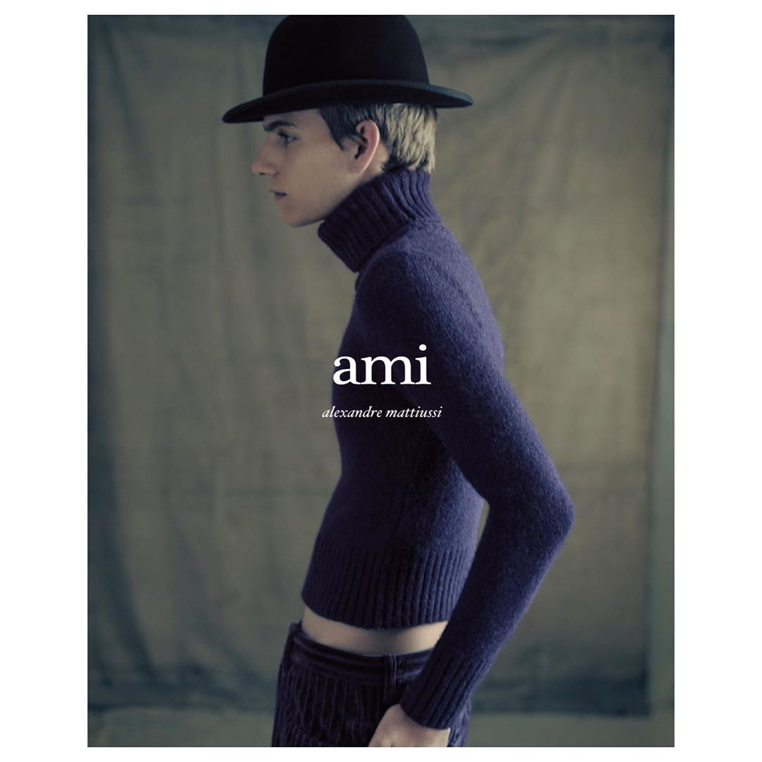 Fashion portrait of a blonde-haired person wearing a dark turtleneck sweater, dark pants, and a black bowler hat against a grey background.