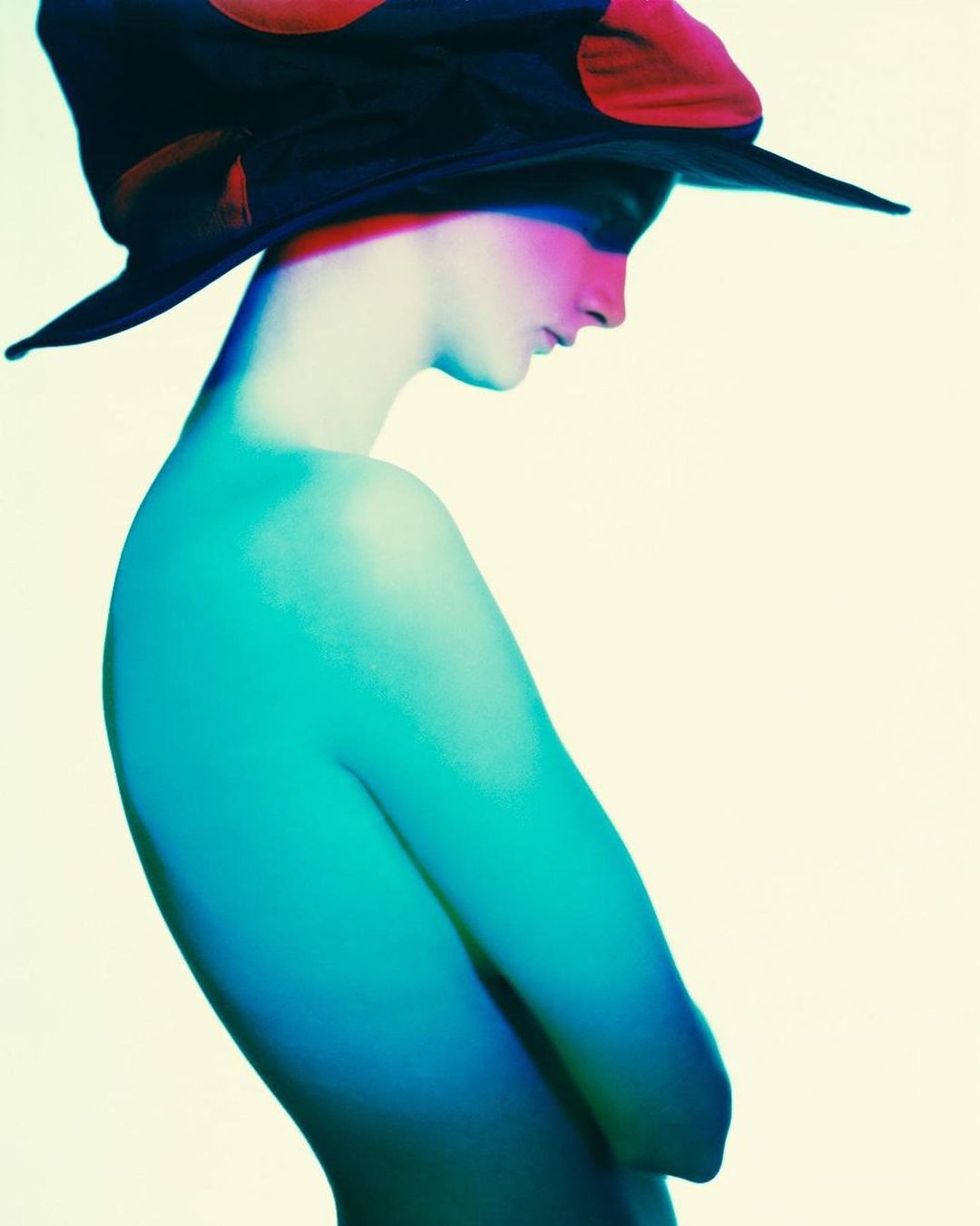 Striking portrait of a fair-skinned person wearing a dark hat with red spots, illuminated by vivid pink and teal lighting against a pale backdrop.