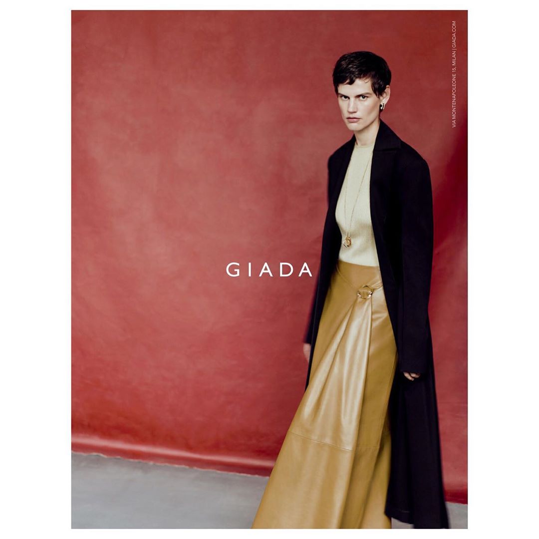 Model with short dark hair wearing a light turtleneck, tan skirt, and black coat against a textured red backdrop.