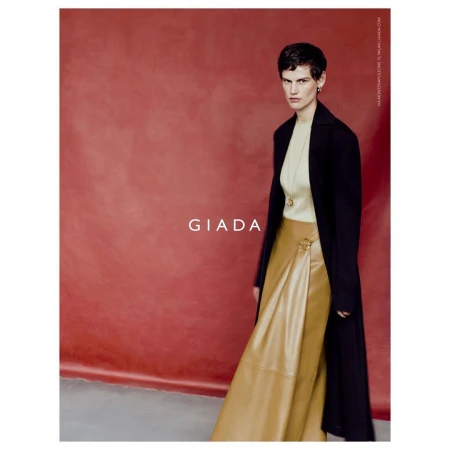 Model with short dark hair wearing a light turtleneck, tan skirt, and black coat against a textured red backdrop.