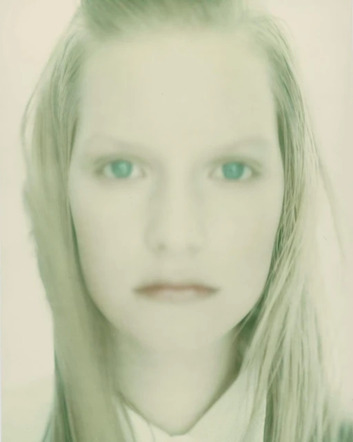 Close-up portrait of a fair-skinned adult female with blonde hair, light-colored clothing, and striking blue eyes, presented in soft, diffused light.