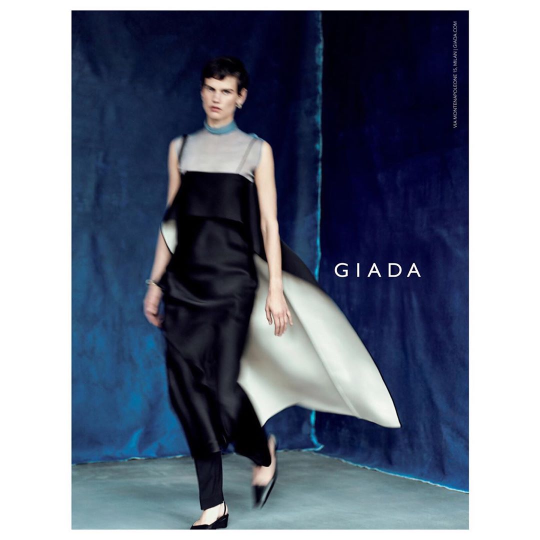 Model in black and grey layered dress with flowing cape walking against a textured blue backdrop