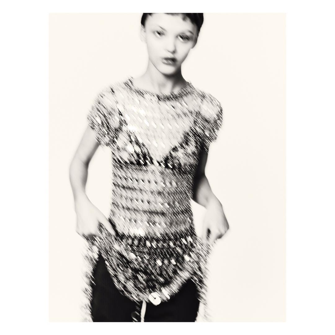 A young woman in a beaded top poses in a studio. The focus is soft and lends the image an artistic feel. The background is plain.