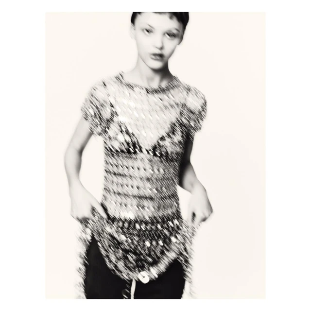A young woman in a beaded top poses in a studio. The focus is soft and lends the image an artistic feel. The background is plain.