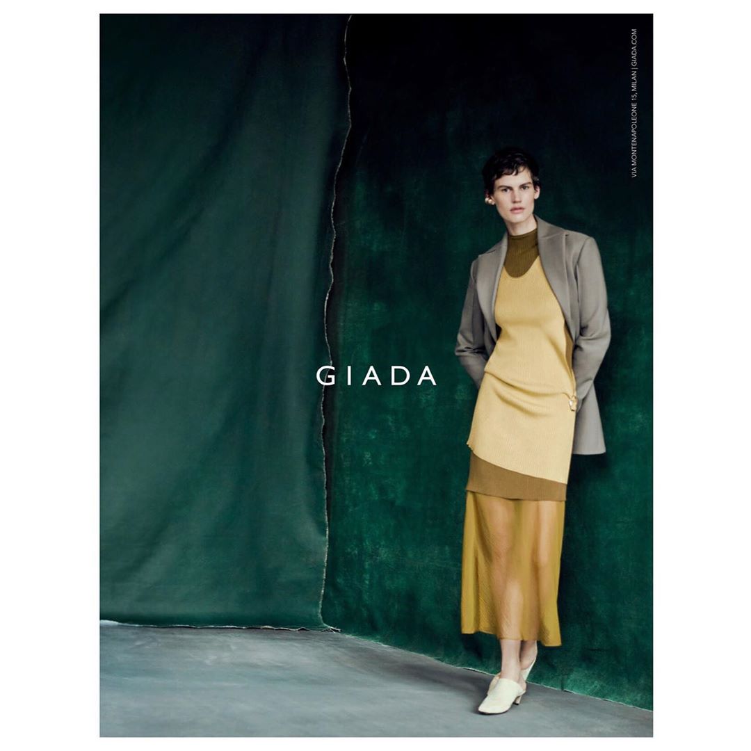 A woman poses in a studio wearing a two-tone tan and beige layered dress and grey jacket by Giada, in a fashion editorial shot.
