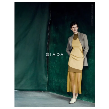 A woman poses in a studio wearing a two-tone tan and beige layered dress and grey jacket by Giada, in a fashion editorial shot.