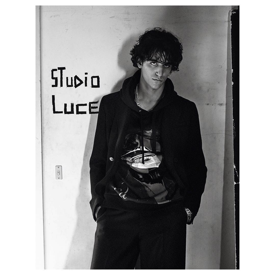 Portrait of a young adult male in a black hoodie with a graphic, standing indoors in front of a white wall with graffiti. Monochrome.