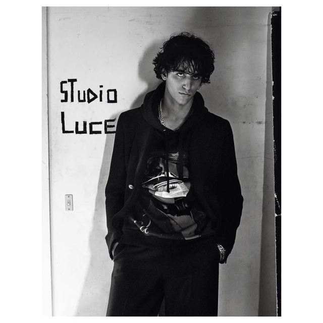 Portrait of a young adult male in a black hoodie with a graphic, standing indoors in front of a white wall with graffiti. Monochrome.
