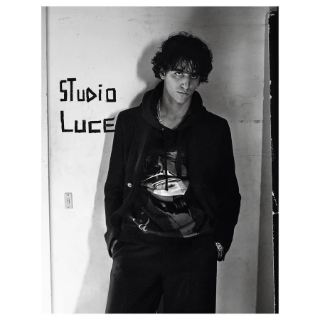 Portrait of a young adult male in a black hoodie with a graphic, standing indoors in front of a white wall with graffiti. Monochrome.