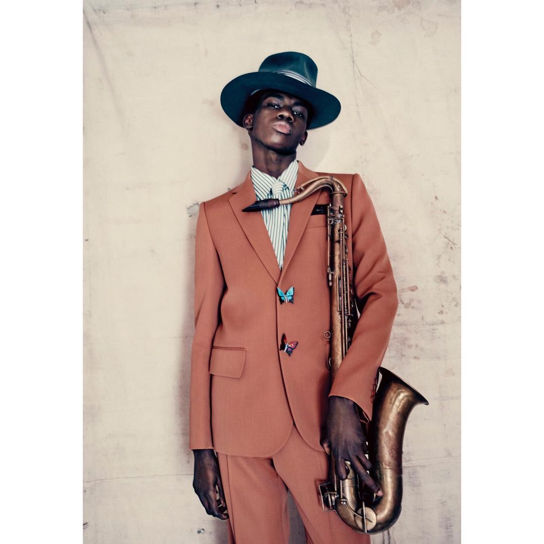 An African American man in a salmon suit and green hat poses with a saxophone in front of a neutral wall, creating a stylish musical portrait.