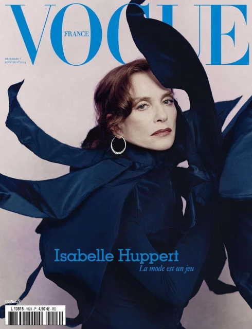Vogue France magazine cover featuring adult female Isabelle Huppert in navy blue, with red hair and hoop earrings, on a light pink background.