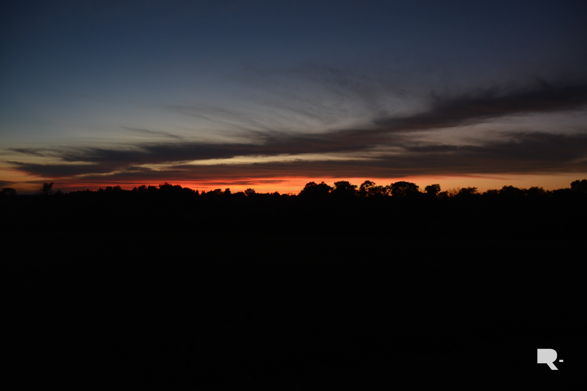 Silhouetted treeline beneath a dramatic sunset sky with vibrant orange glow on the horizon and dark clouds above.