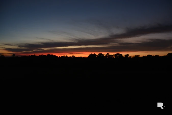 Silhouetted treeline beneath a dramatic sunset sky with vibrant orange glow on the horizon and dark clouds above.