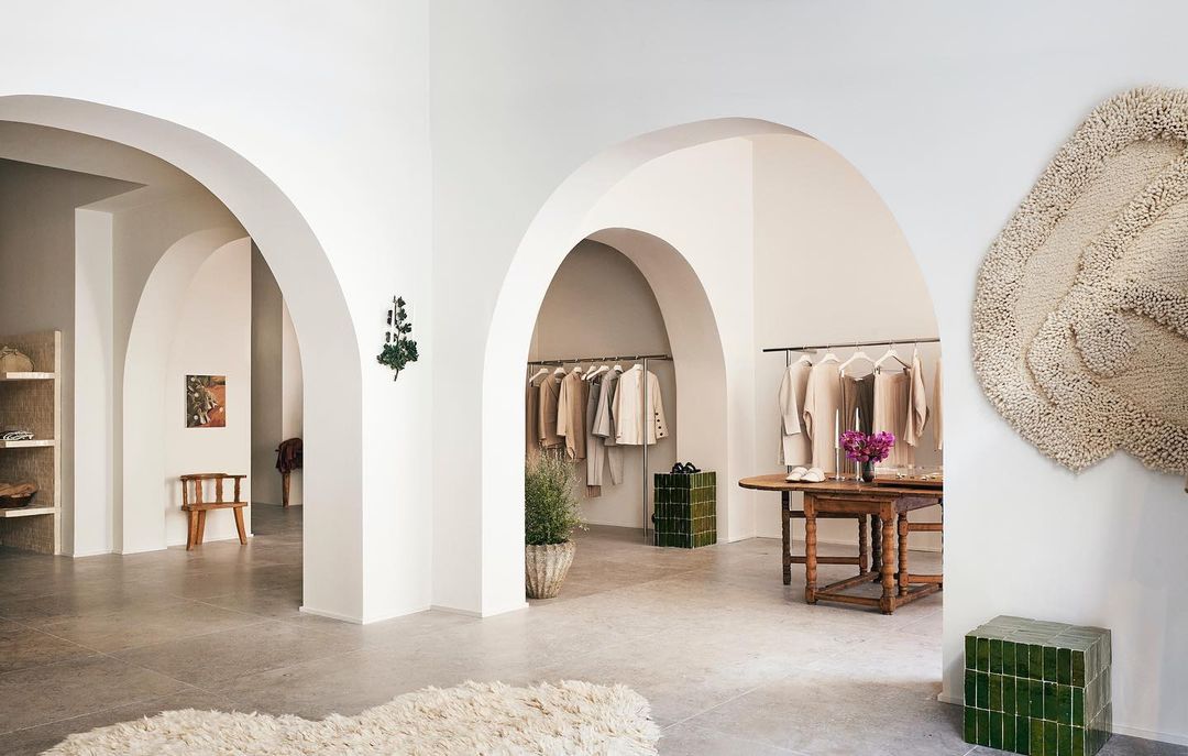 Clothing displayed in an airy boutique with arched doorways, neutral color palette, potted plants, and decorative wall hanging.