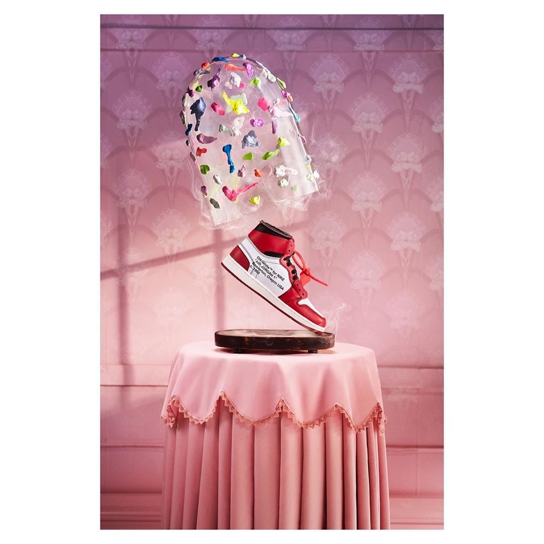 Floating red and white Nike Air Jordan shoe above a pink ruffled table and against pink patterned wallpaper with a colorful veil.