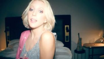 Portrait of a blond adult female singing in an indoor bedroom setting, wearing a tank top and cardigan, with a guitar in the background.