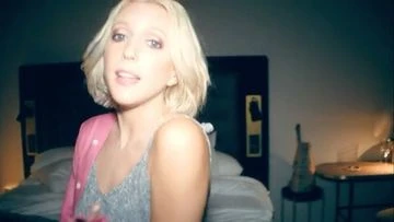 Portrait of a blond adult female singing in an indoor bedroom setting, wearing a tank top and cardigan, with a guitar in the background.