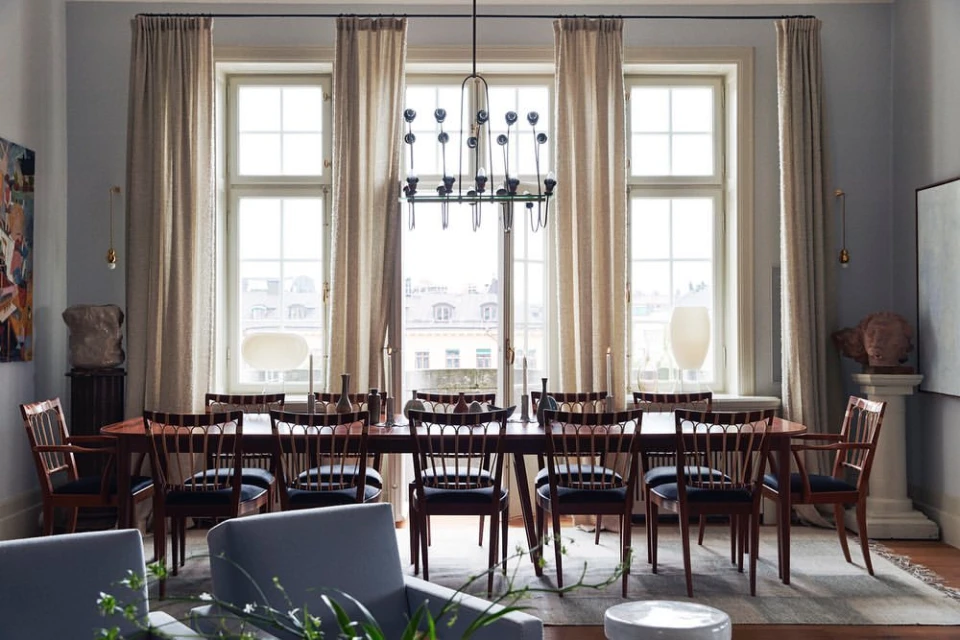 Elegant dining room featuring a long wood table with chairs, a modern chandelier, and large windows with light-colored curtains.