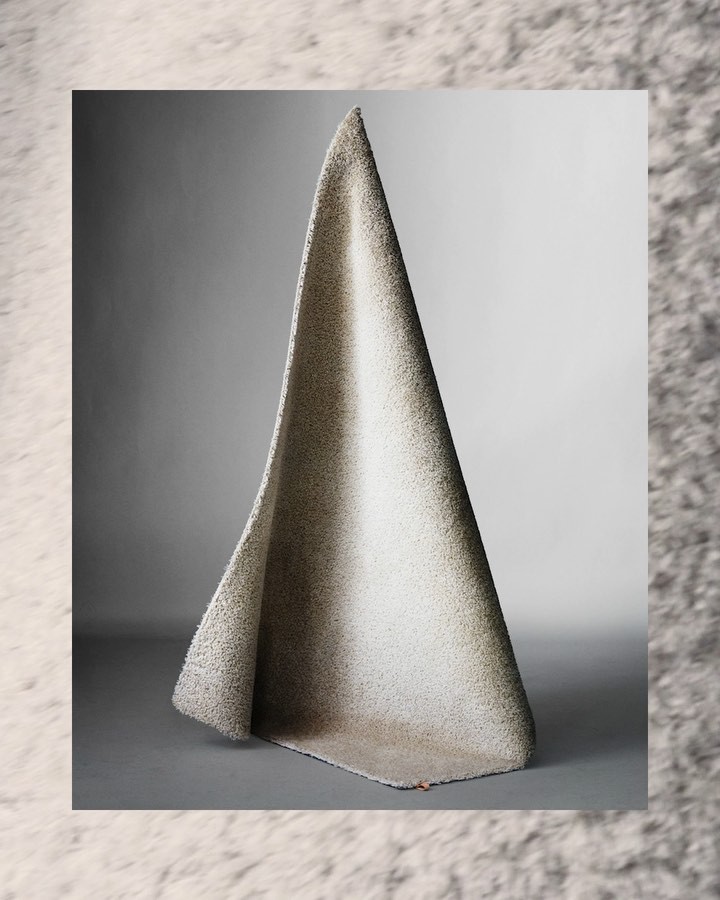 A speckled cream and brown rug is draped into a peaked triangular shape, artfully posed in a simple gray studio setting.