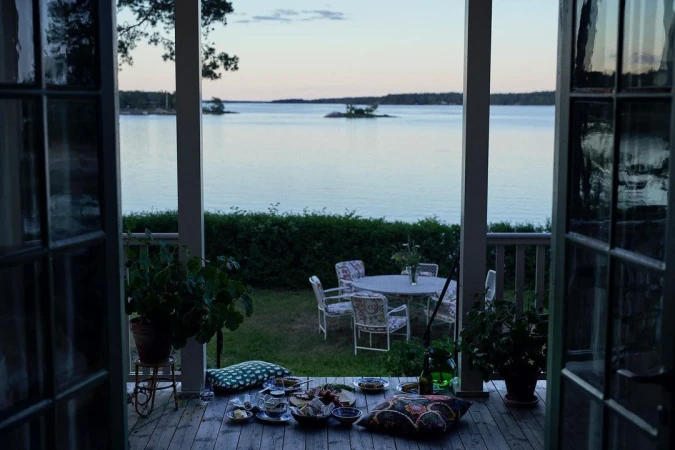 Outdoor dining area overlooking a tranquil lake at dusk. Table setting with tableware and cushions on a wooden deck next to plants.