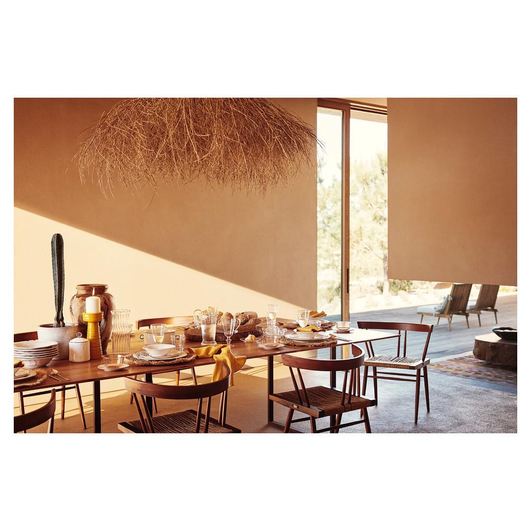 Dining table set with plates, glasses, and decorative items under a straw light fixture in a brightly lit room with outdoor views.
