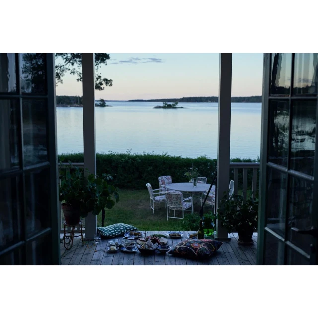 Outdoor dining scene on a porch overlooking a serene lake at dusk, featuring a set table, seating, cushions, and potted plants.