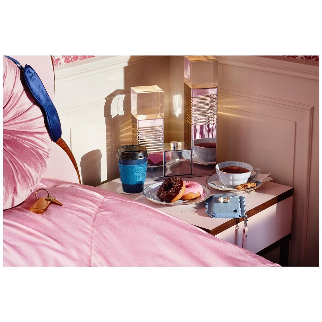 Still life of bedroom items: coffee cup, donuts, sleep mask, and keys, arranged on a nightstand with pastel pink bedding.