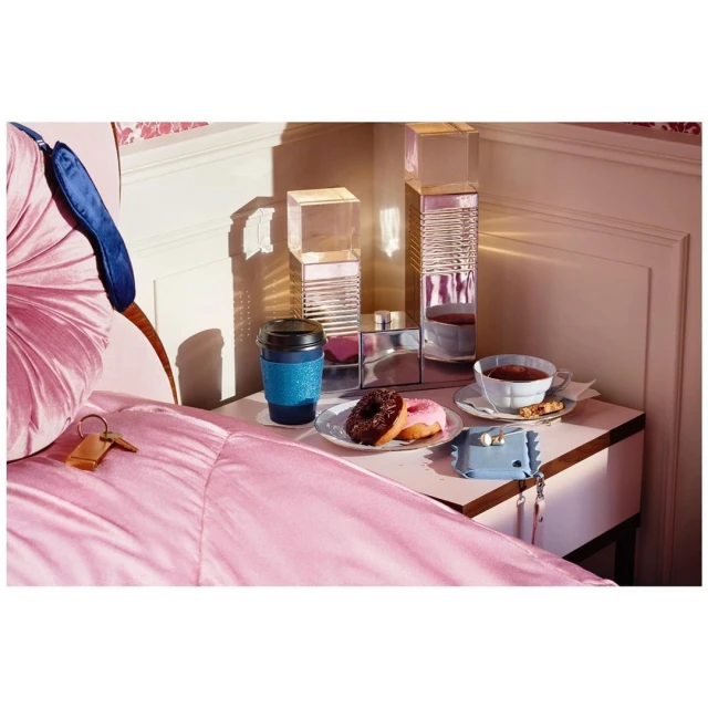 Still life of bedroom items: coffee cup, donuts, sleep mask, and keys, arranged on a nightstand with pastel pink bedding.