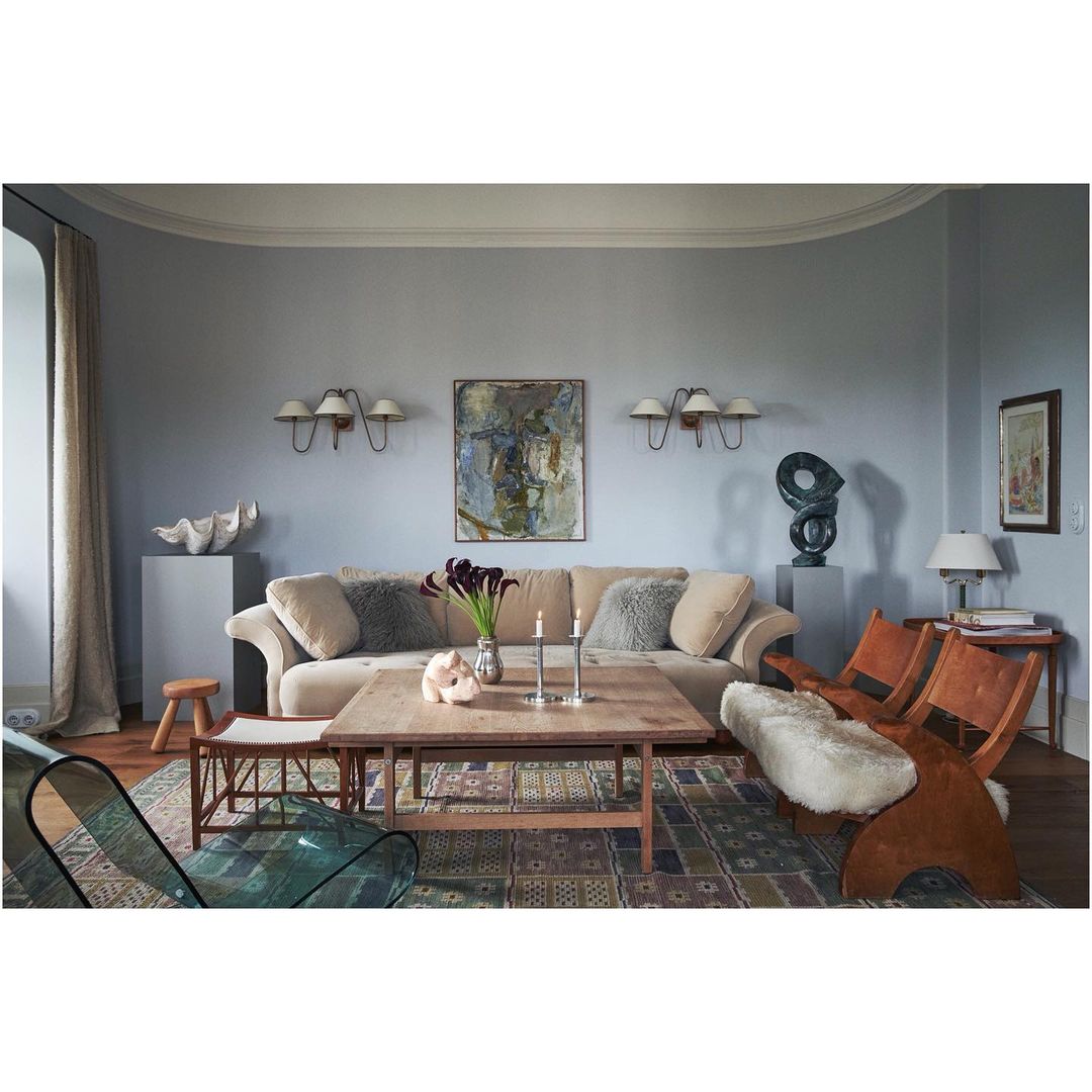 Living room interior featuring a beige couch with decorative pillows, wooden coffee table with floral arrangement, and two wooden chairs.