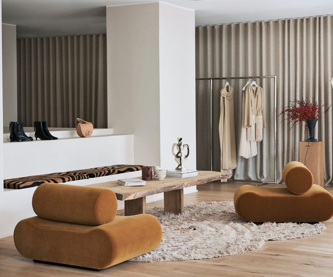 Interior design of a minimalist living room featuring a wooden coffee table, armchairs, a clothing rack, and neutral color palette.