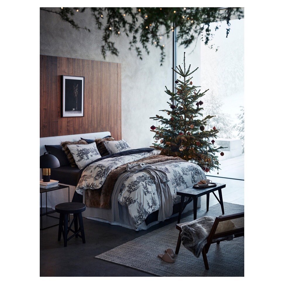 Cozy bedroom interior with a decorated Christmas tree near a large window. Bed with patterned duvet, wooden accents and rug.