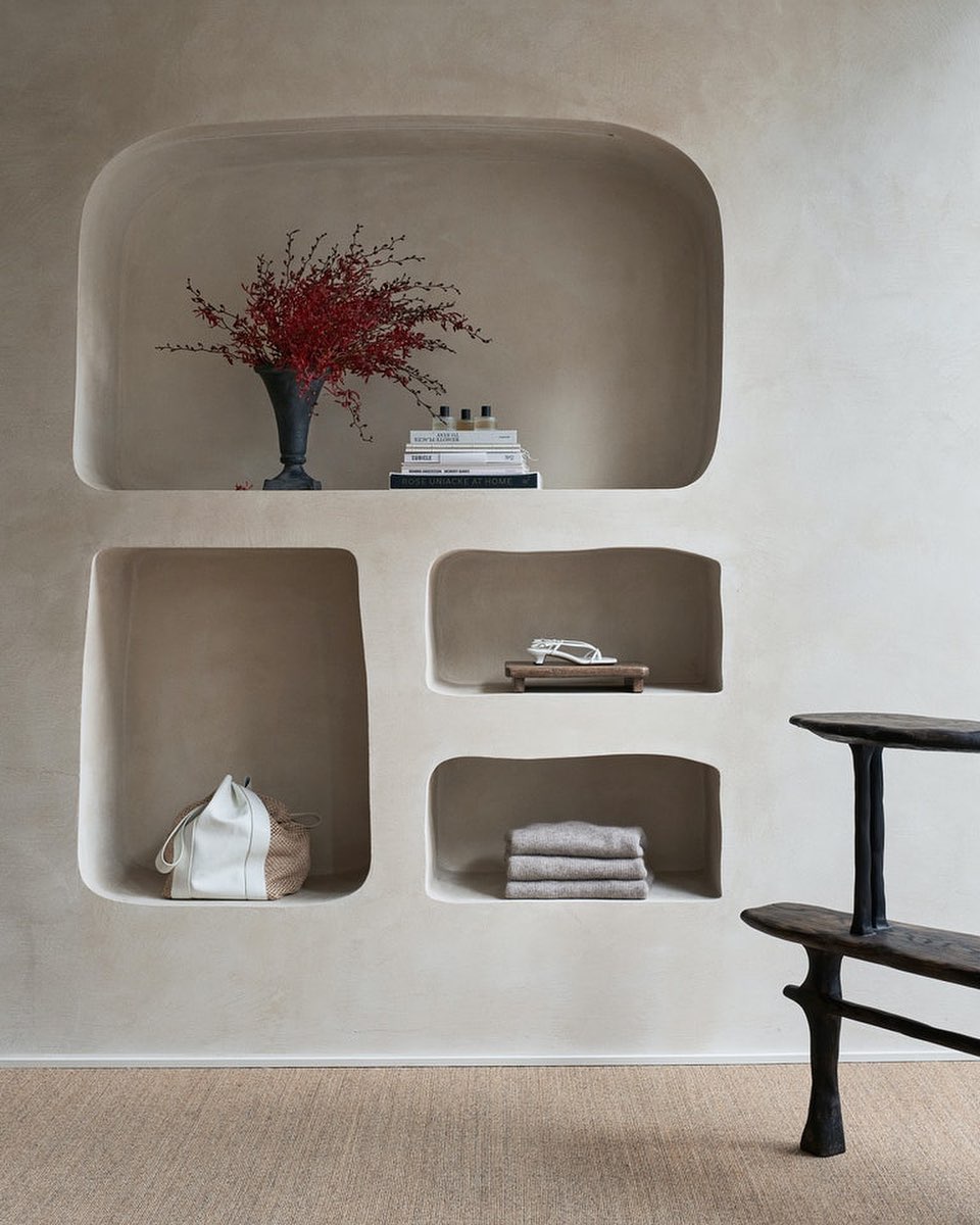 Minimalist interior featuring recessed shelving with red floral arrangement, stacked books, self care items, and neutral-toned textiles.
