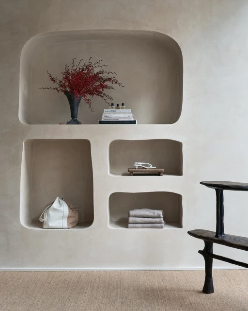 Minimalist interior featuring recessed shelving with red floral arrangement, stacked books, self care items, and neutral-toned textiles.