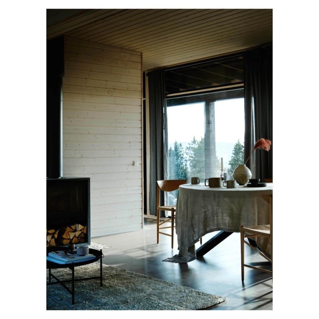 A bright and modern interior with a round dining table, chairs, fireplace, wooden walls and ceilings, and large windows offering a forest view.