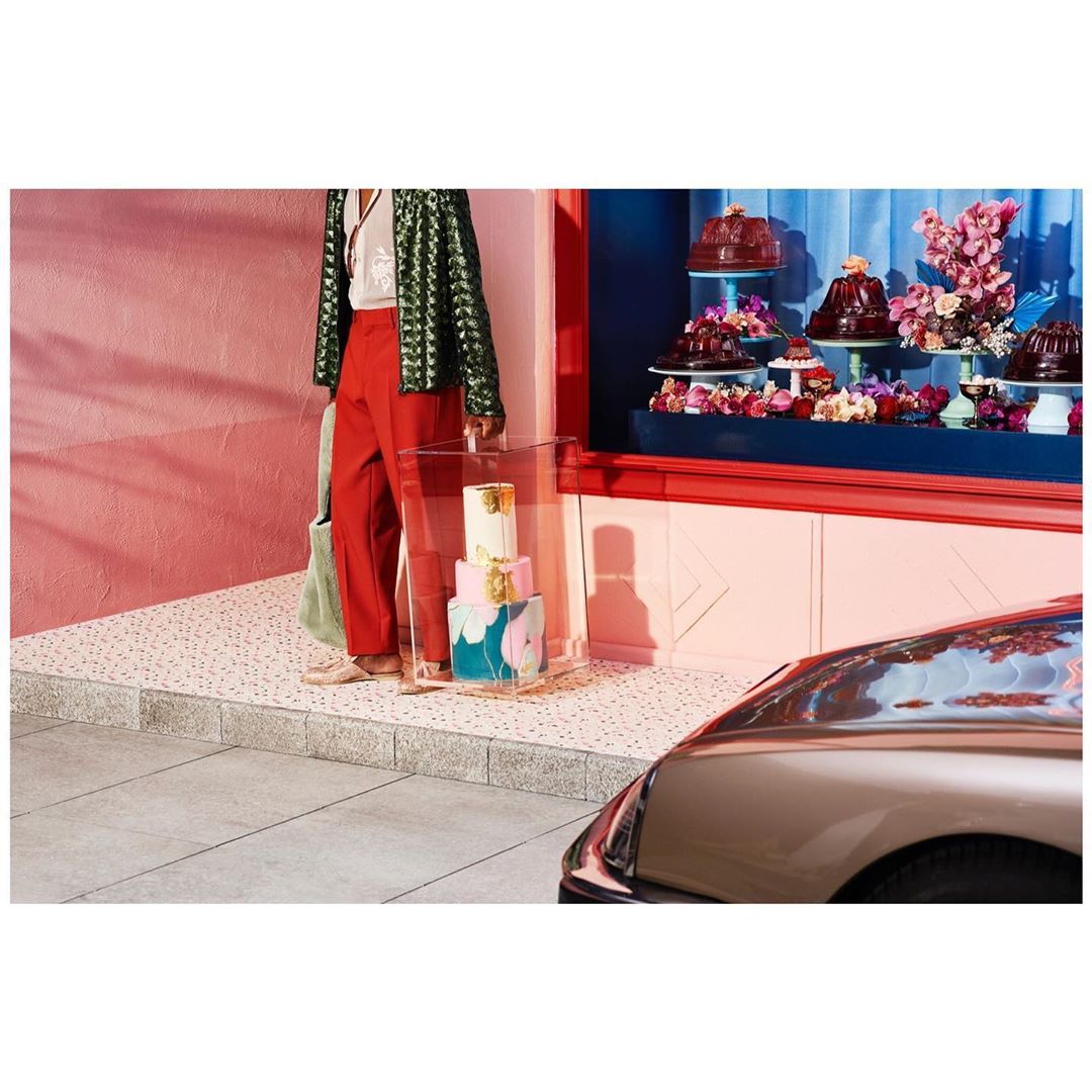 Woman in red pants stands near a window display with cakes and flowers, next to a parked car on a city street with speckled sidewalk.