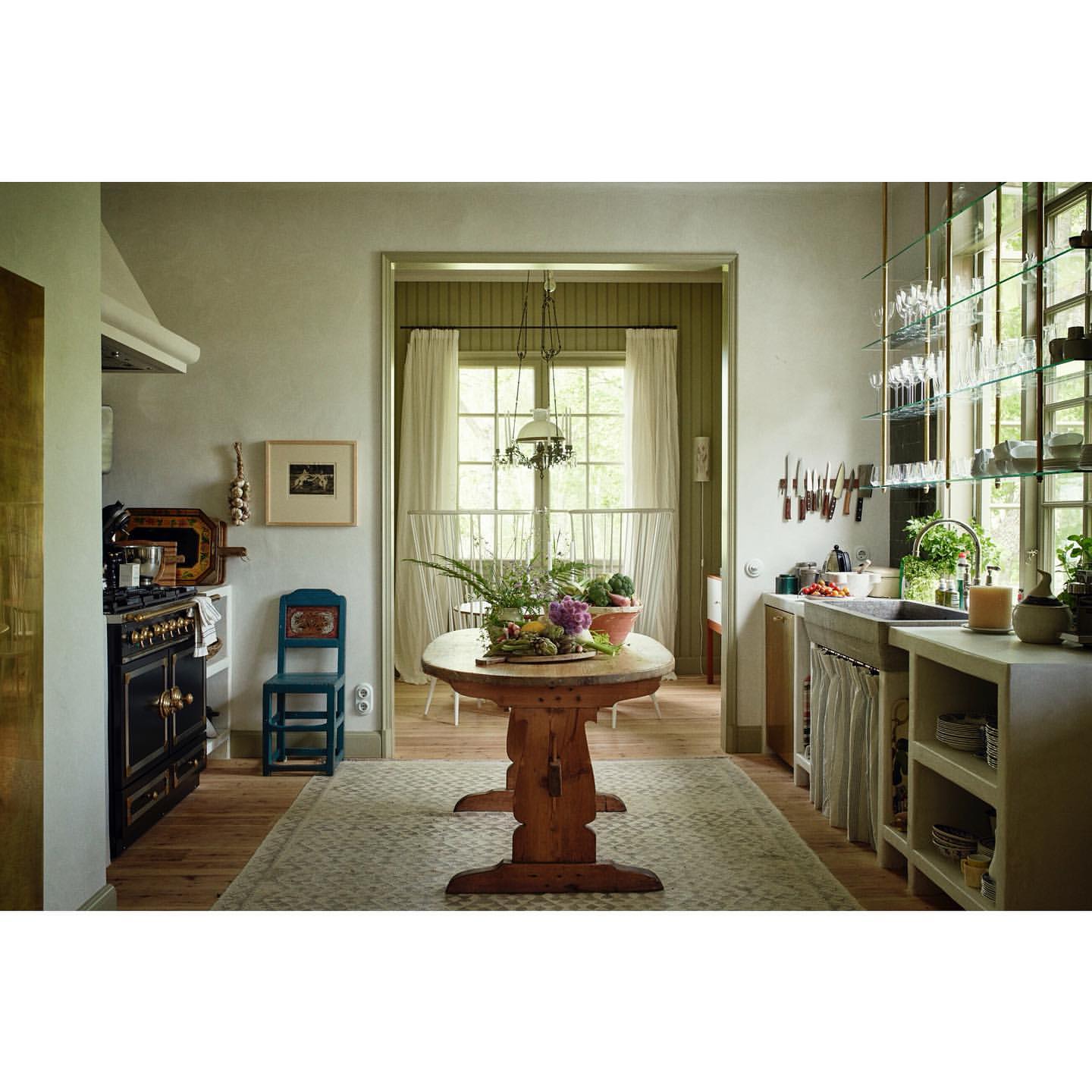 Interior view of a rustic kitchen featuring a wooden table with produce, a vintage stove, and natural light from a window.
