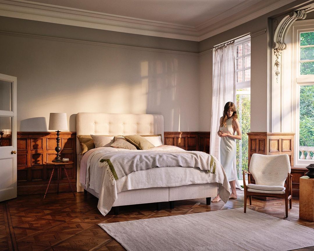 A woman in a light dress stands in a sunlit bedroom with a large bed, wood floors, and sheer curtains.