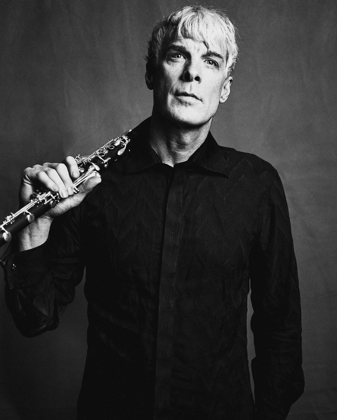Portrait of a fair-skinned adult male with short blonde hair in a black shirt holding a woodwind instrument against a neutral background.